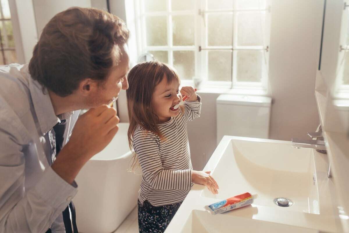 Father and daughter brushing teeth together at bathroom sink during dental hygiene routine.