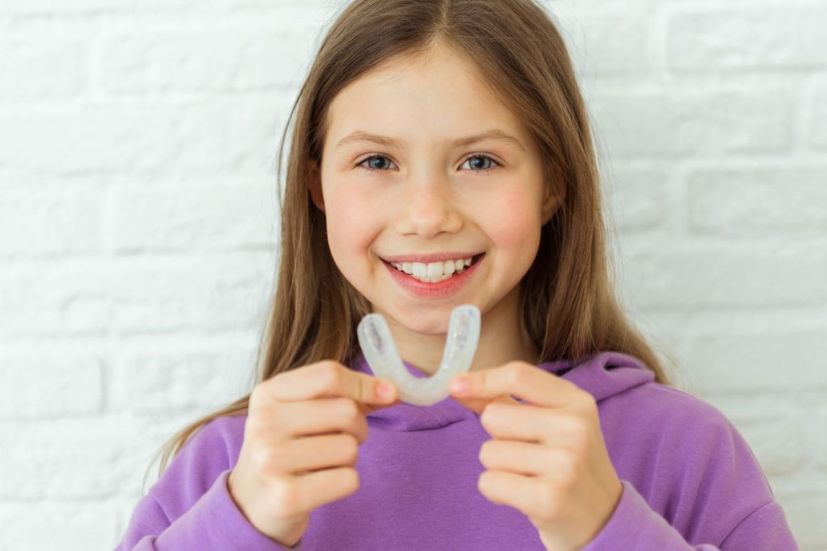 Happy girl in purple shirt holding clear mouthguard and smiling at camera.